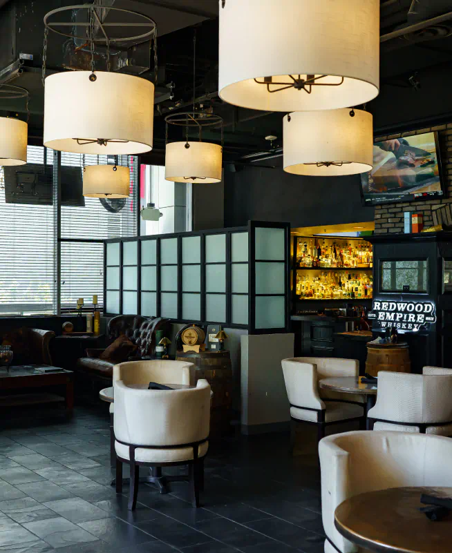 Interior of a lobby with large lights suspended from the ceiling. Small tables are in the foreground and behind a divider in the background there is an entrance to a bar.