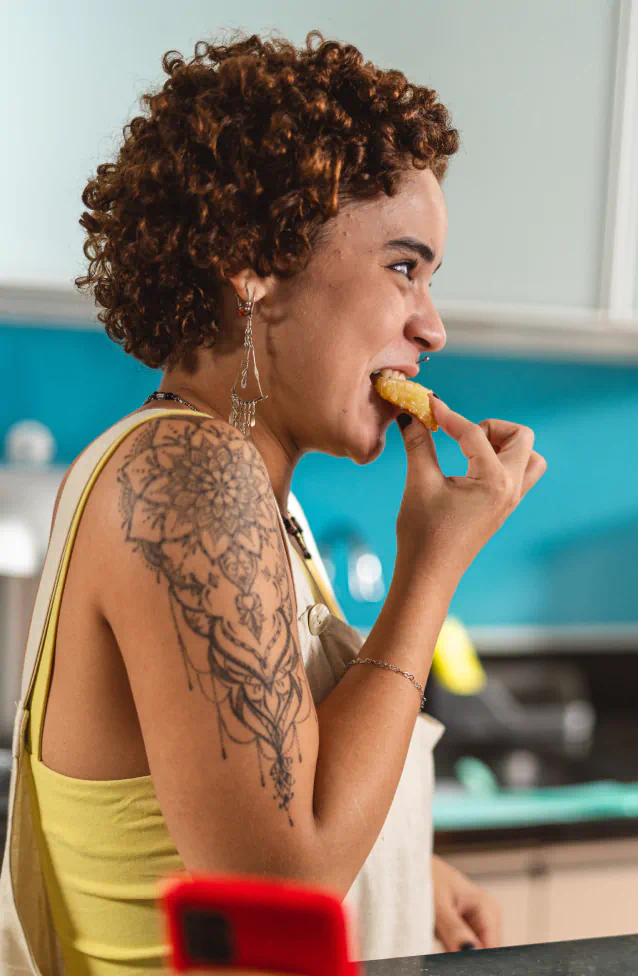 Woman with curly hair and floral arm tattoo eating a snack in a kitchen.