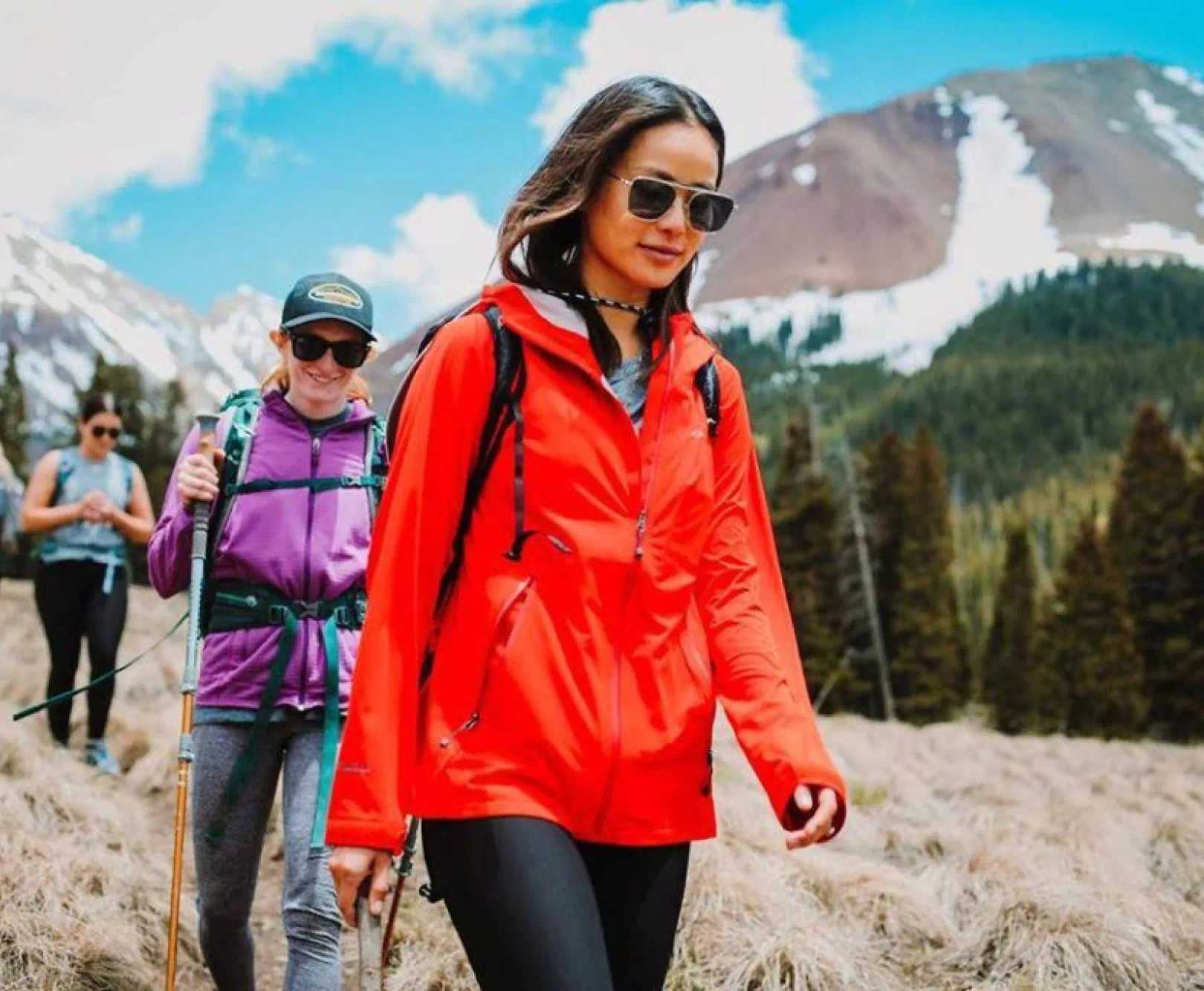 Hikers walking on a grassy trail with mountains and trees in the background.