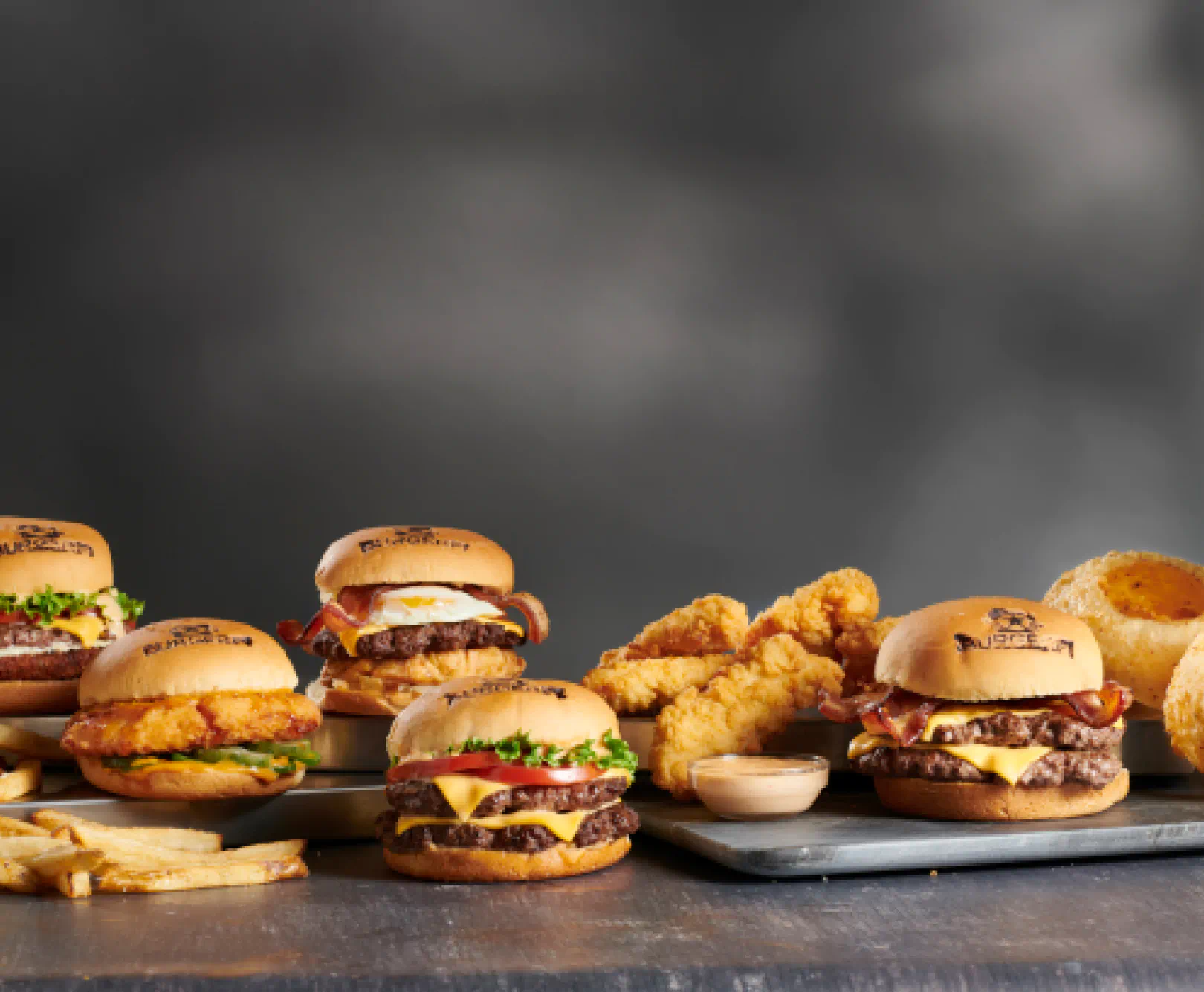 Several burgers, fried chicken tenders, an onion ring, and fries arranged on a gray surface.