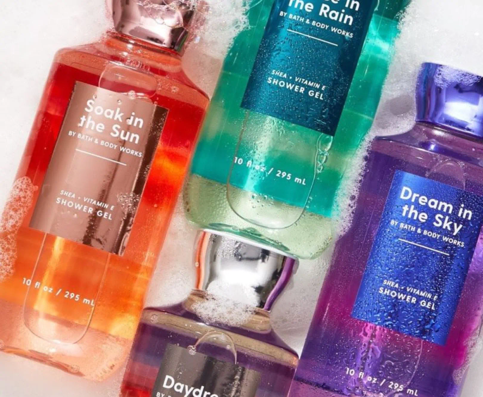 Four different colored Bath and Body Works shower gel bottles floating in soapy water, including "soak in the sun" "daydream" "dream in the sky" and "dance in the rain"