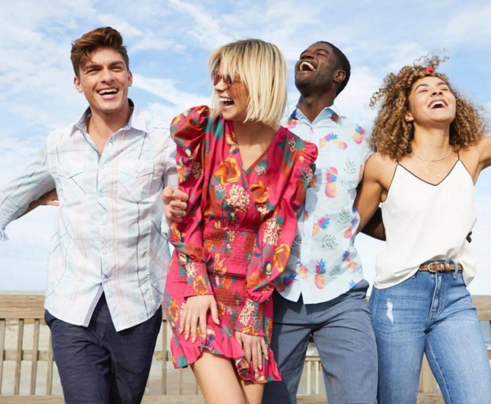 Four people smiling and laughing together outdoors, wearing colorful clothing against a sky background.