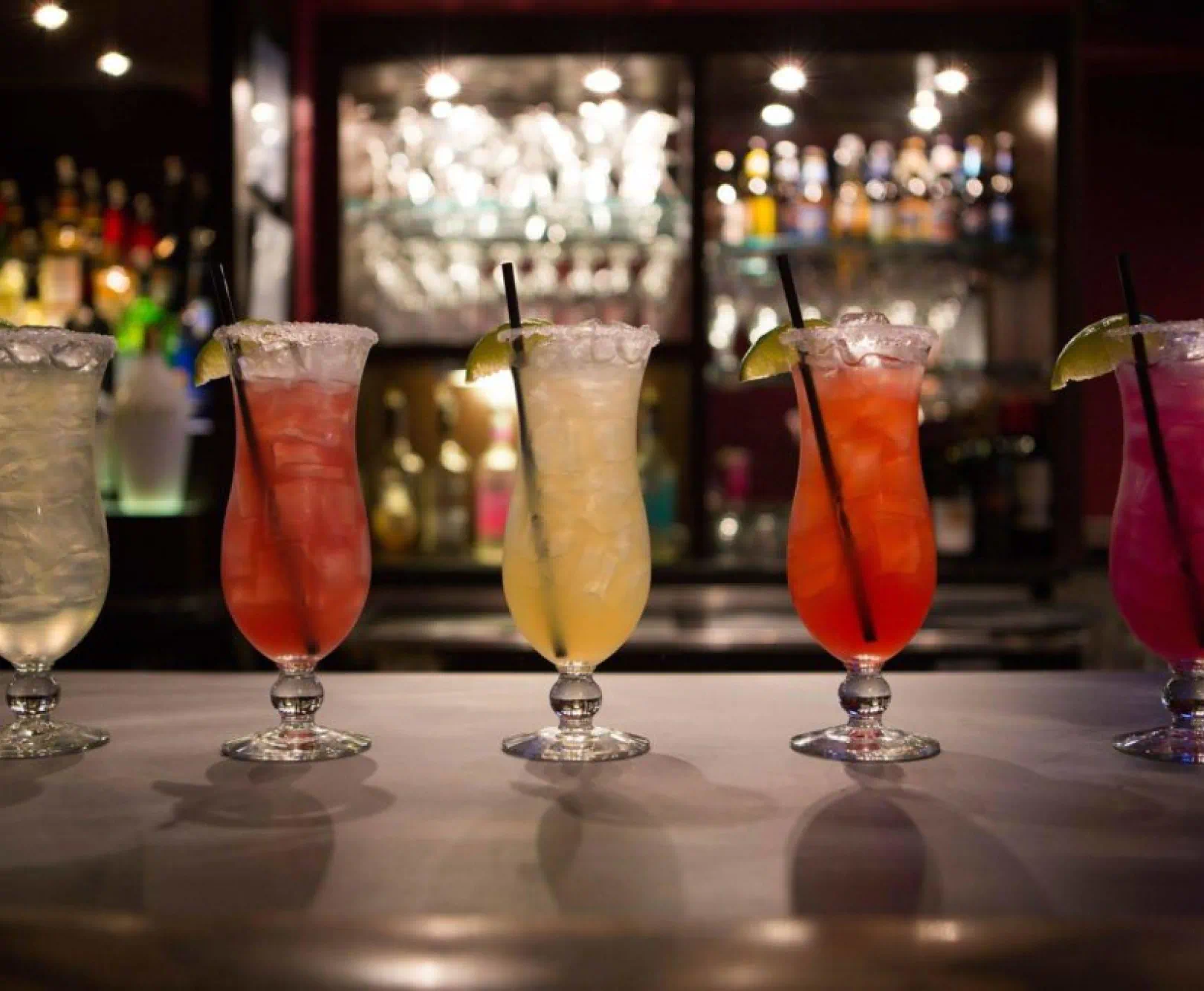 Five colorful cocktails on a bar counter with a backdrop of illuminated liquor bottles.