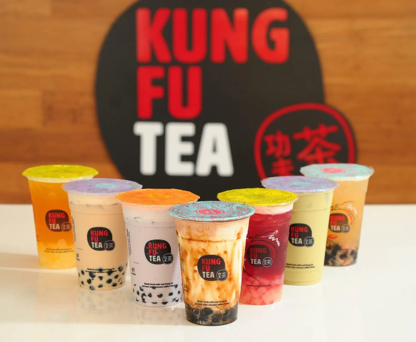 An assortment of eight bubble tea drinks from Kung Fu Tea on a white surface, with a large Kung Fu Tea logo in the background.