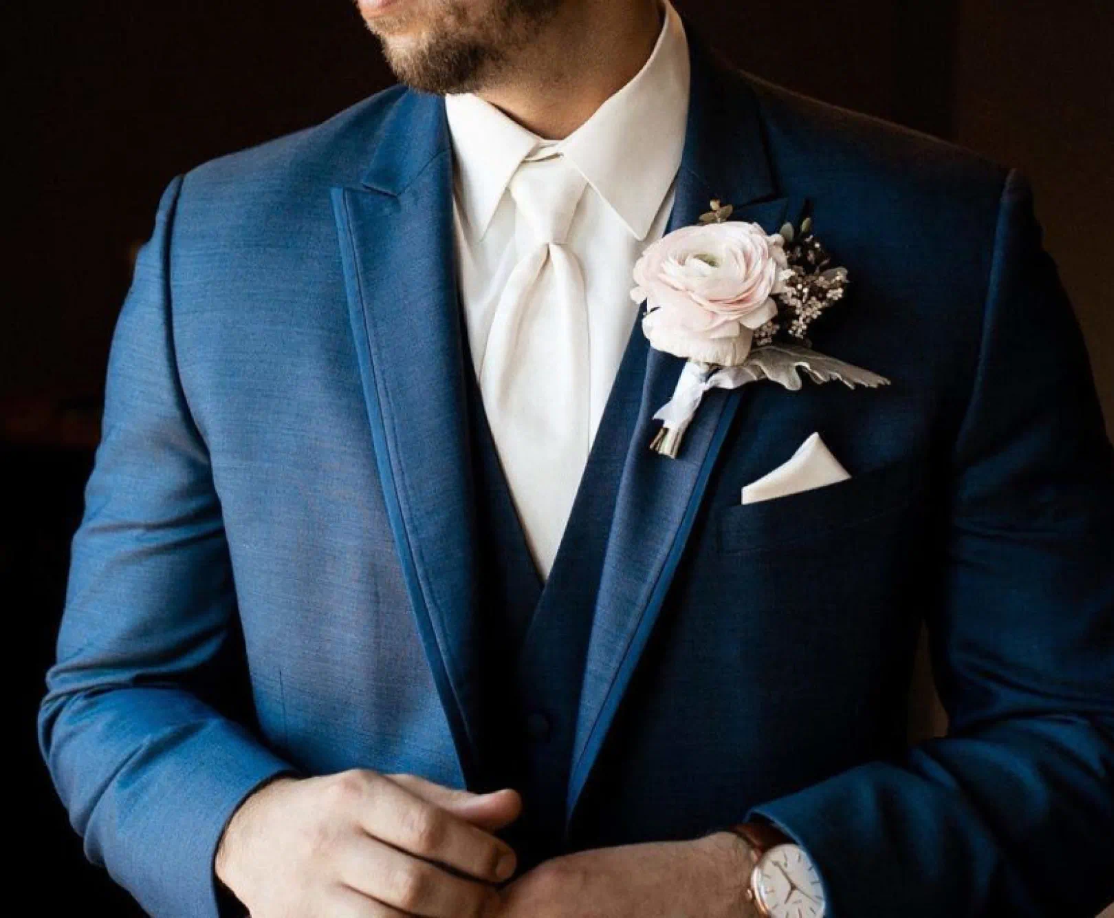 Man in a dark blue suit with a white shirt, tie, and pocket square, wearing a pink rose boutonnière on his left lapel.