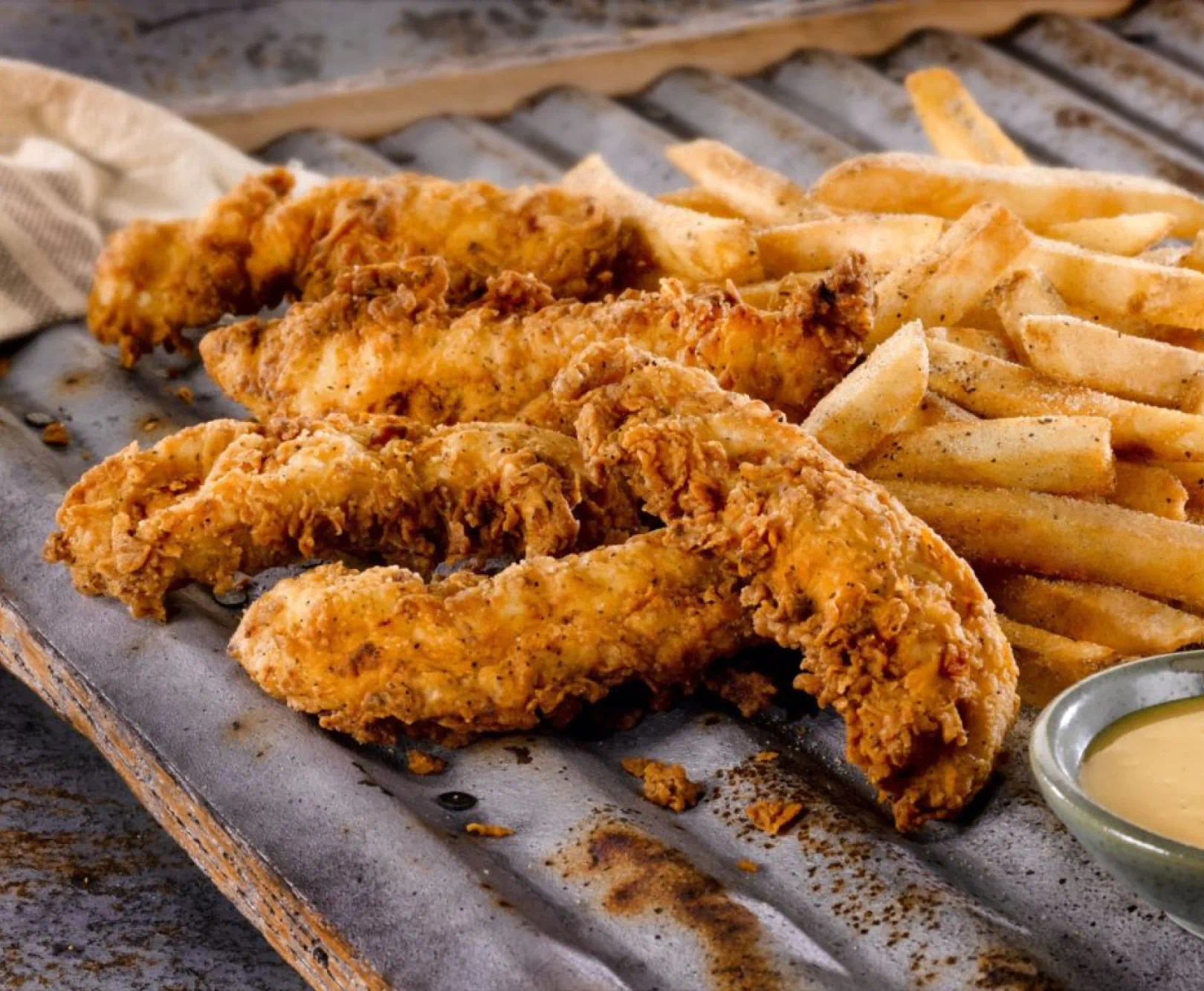 chicken tenders and french fries sit on a currugated metal tray with a dipping sauce partially out of frame