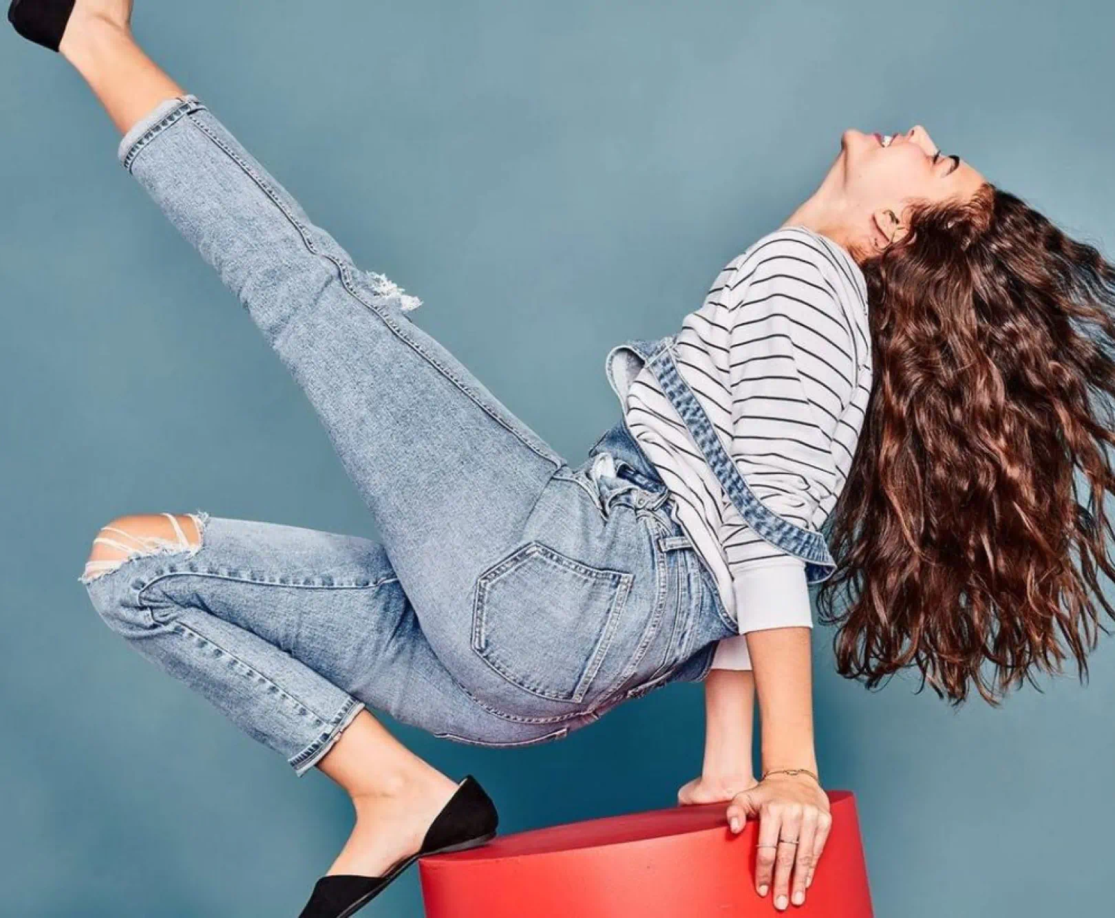 Woman in striped shirt and denim overalls striking a dynamic pose on a red cylinder against a teal background.