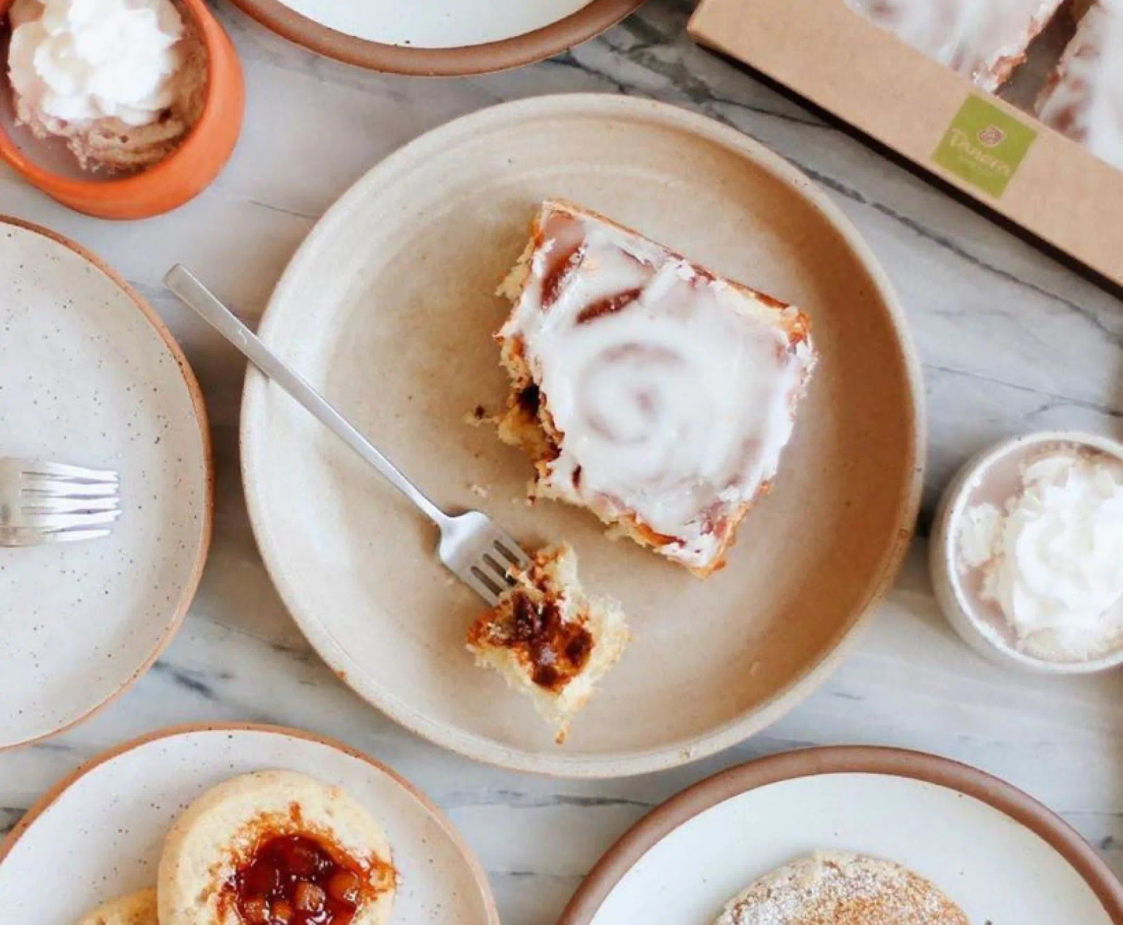 a Panera pastry sits on a tan dish. a piece has been cut off and sits on a fork next to the pastry. various other pastries surround the main dish with a small container of whipped cream beside it.