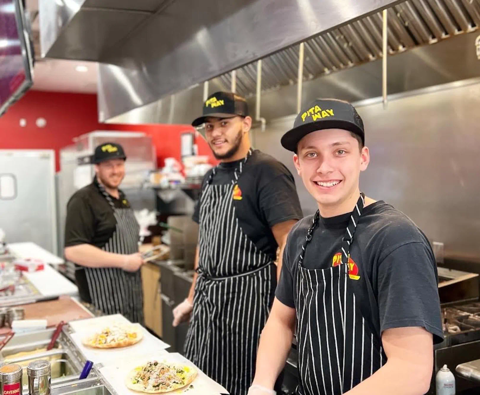 Three people in a commercial kitchen wearing PITA WAY uniforms with black t-shirts, striped aprons, and matching caps.