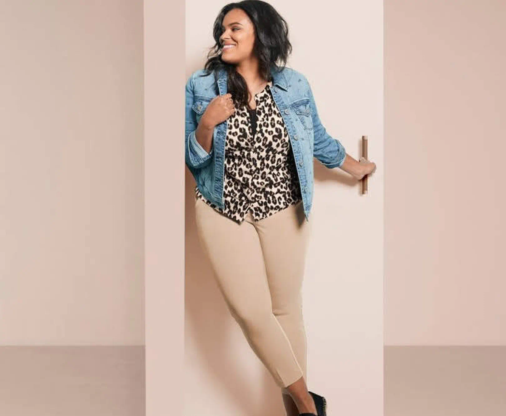 a woman wearing a denim jacket, cheetah print blouse, and khaki pants poses against a beige backdrop