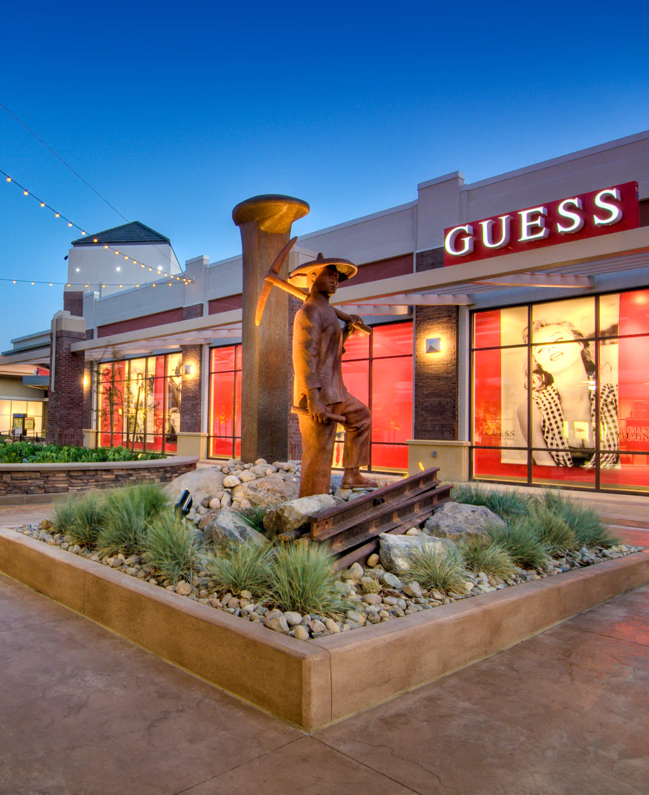 Open air mall with a "Guess" store in the background, and a statue with landscaping in the foreground.