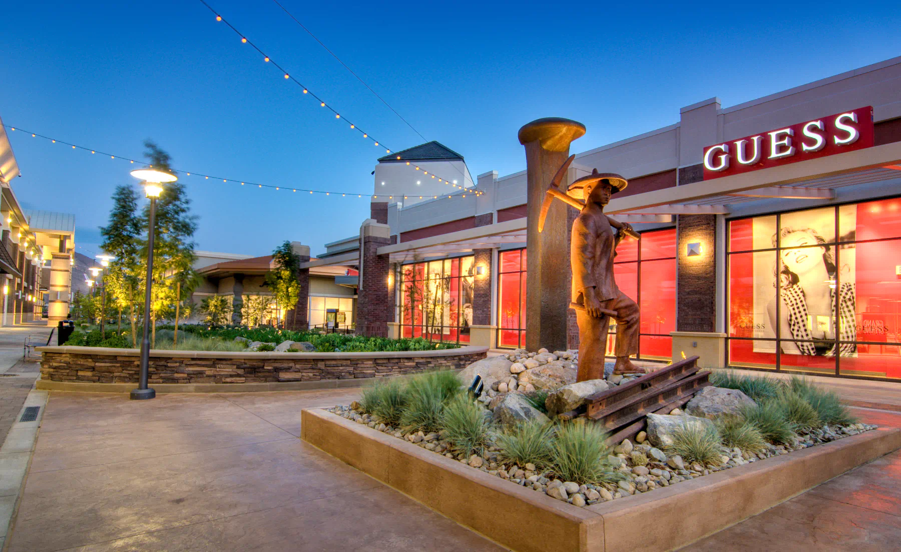 Open air mall with a "Guess" store in the background, and a statue with landscaping in the foreground.