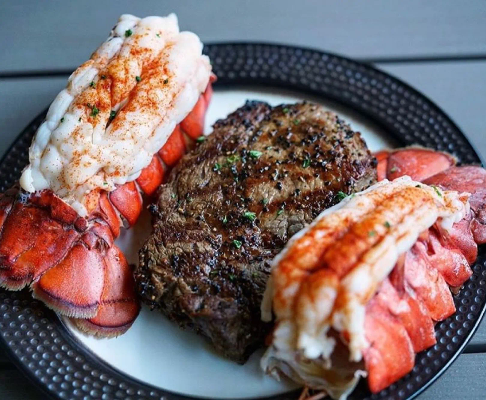 A surf and turf dinner consisting of steak and two lobster tails sits on a black and white plate.