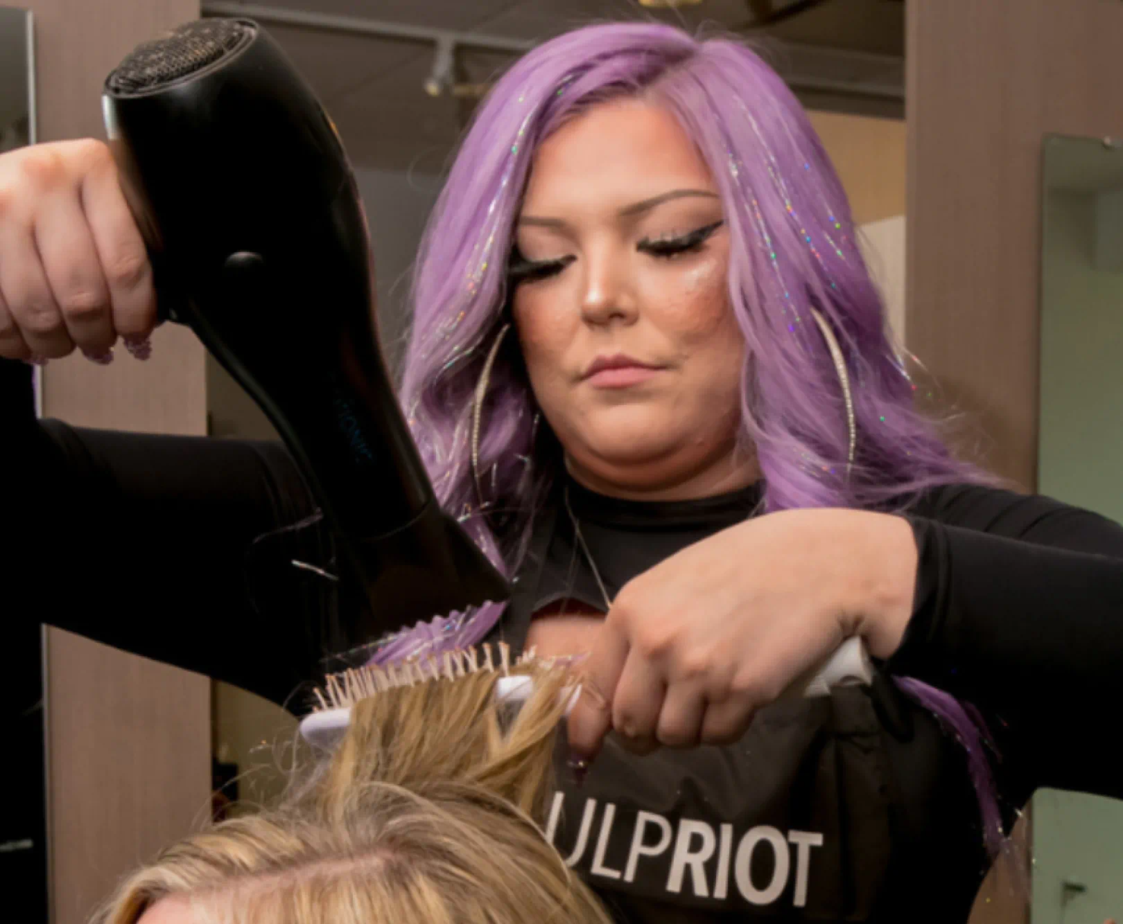 A stylist with purple hair adds volume to a customers blond hair with a hairdryer.