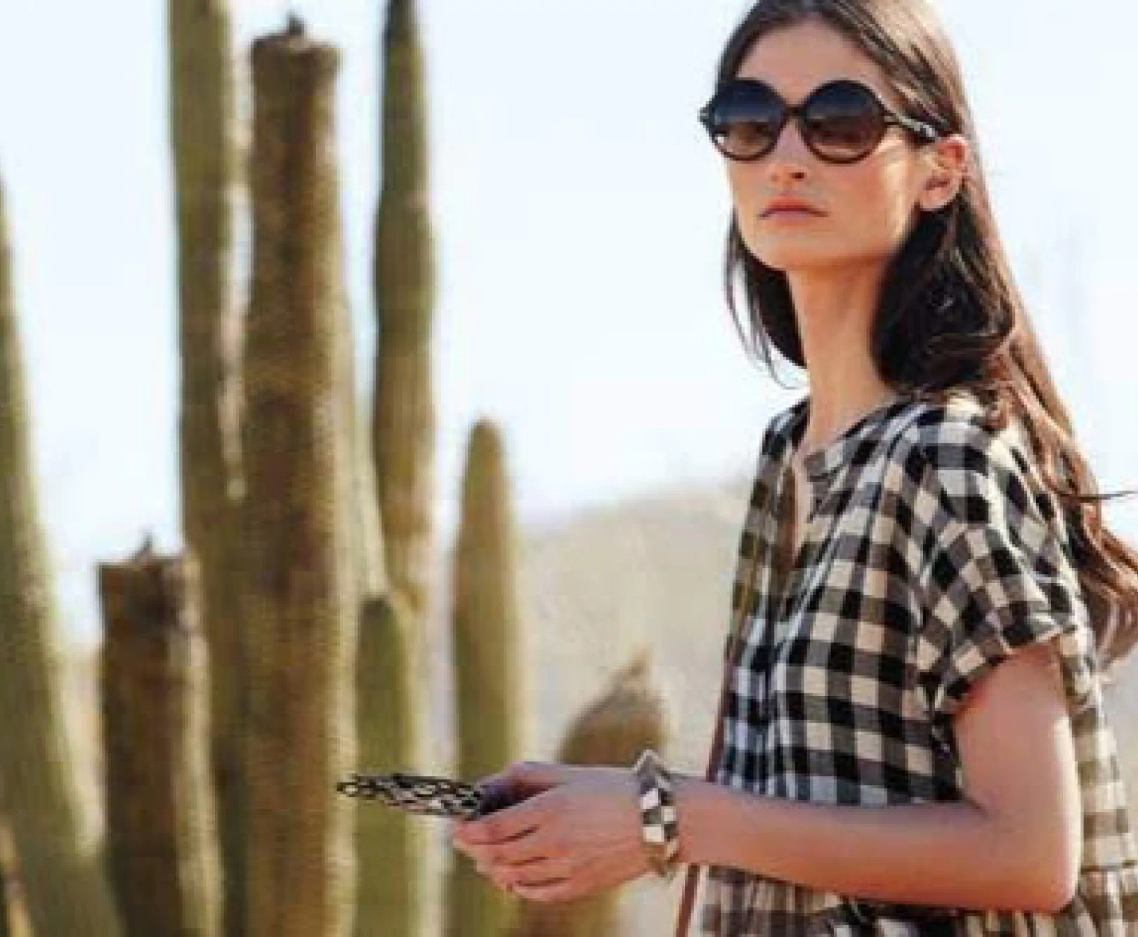 A woman in round, black sunglasses and a checkered top stands in front of a cactus.