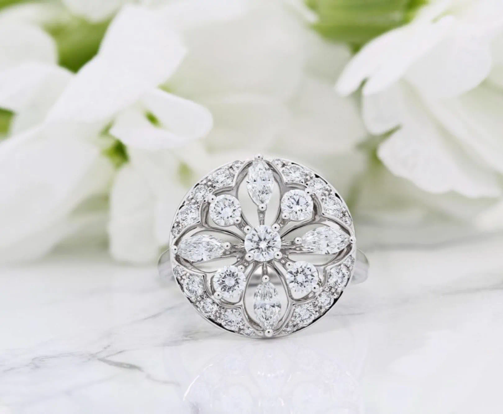 A diamond ring with a floral pattern sit on a white cloth. It's surrounded by white flowers.