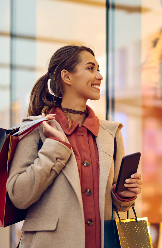 Young woman in a beige wool coat, and rust colored shirt, smiles into shop window, she holds her phone in one hand and shopping bags draped over her shoulder in the other hand.