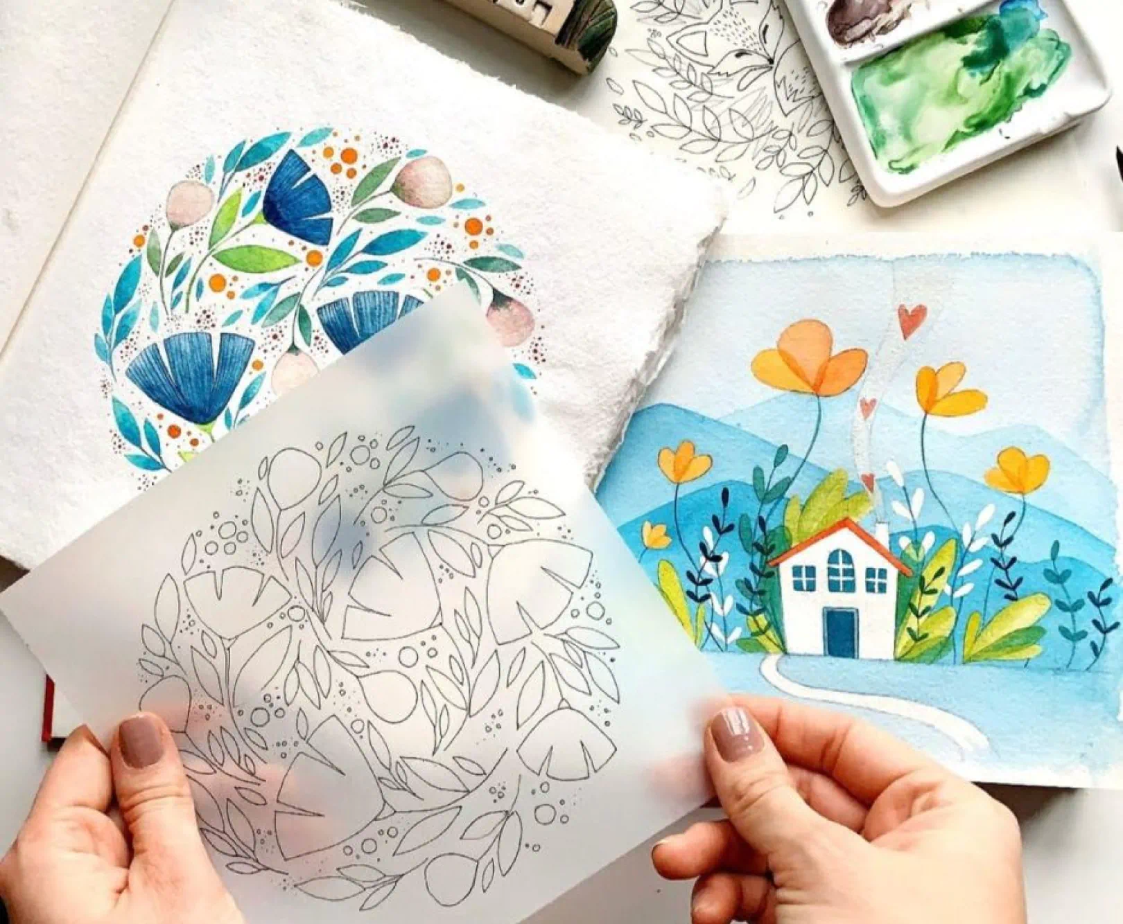 Hands holding a semi-transparent sheet with line drawings of leaves and flowers, surrounded by watercolor artworks and a paint palette.