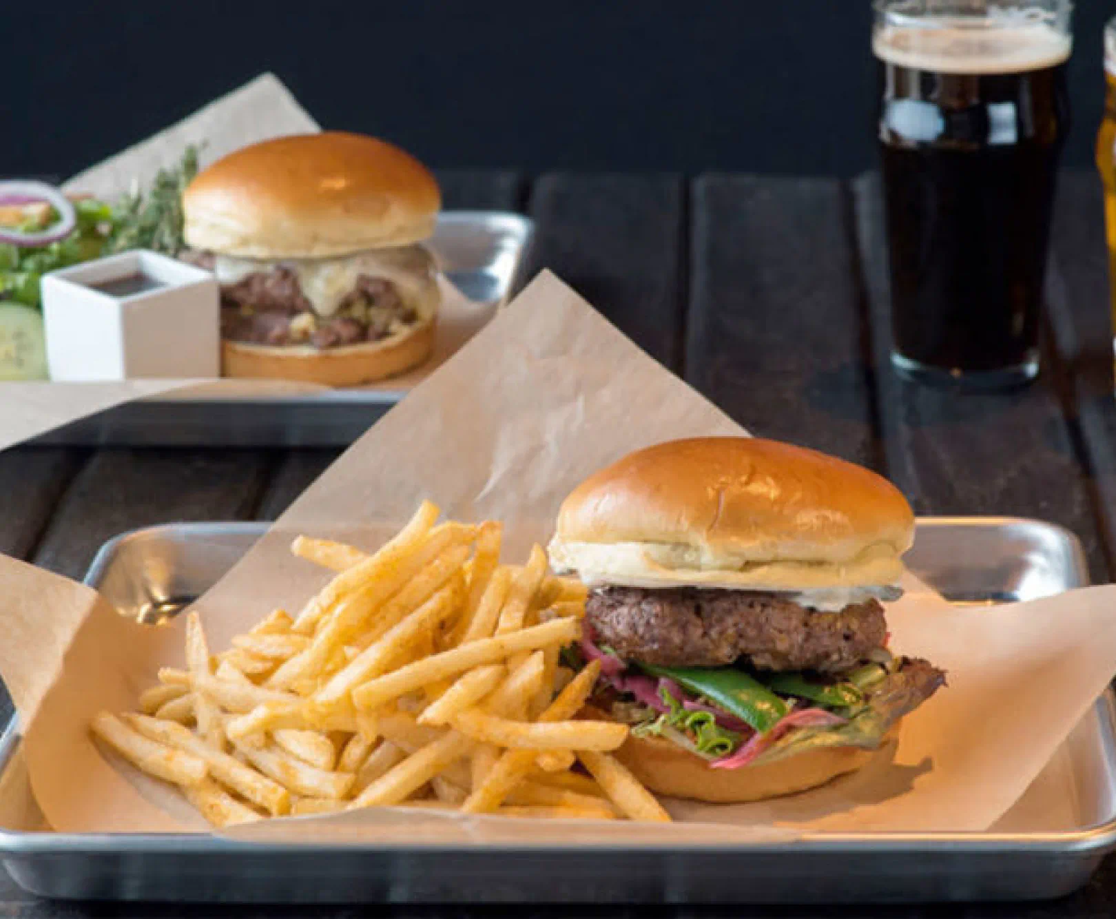 Tray with a burger and fries, with another burger and a glass of dark beer in the background.