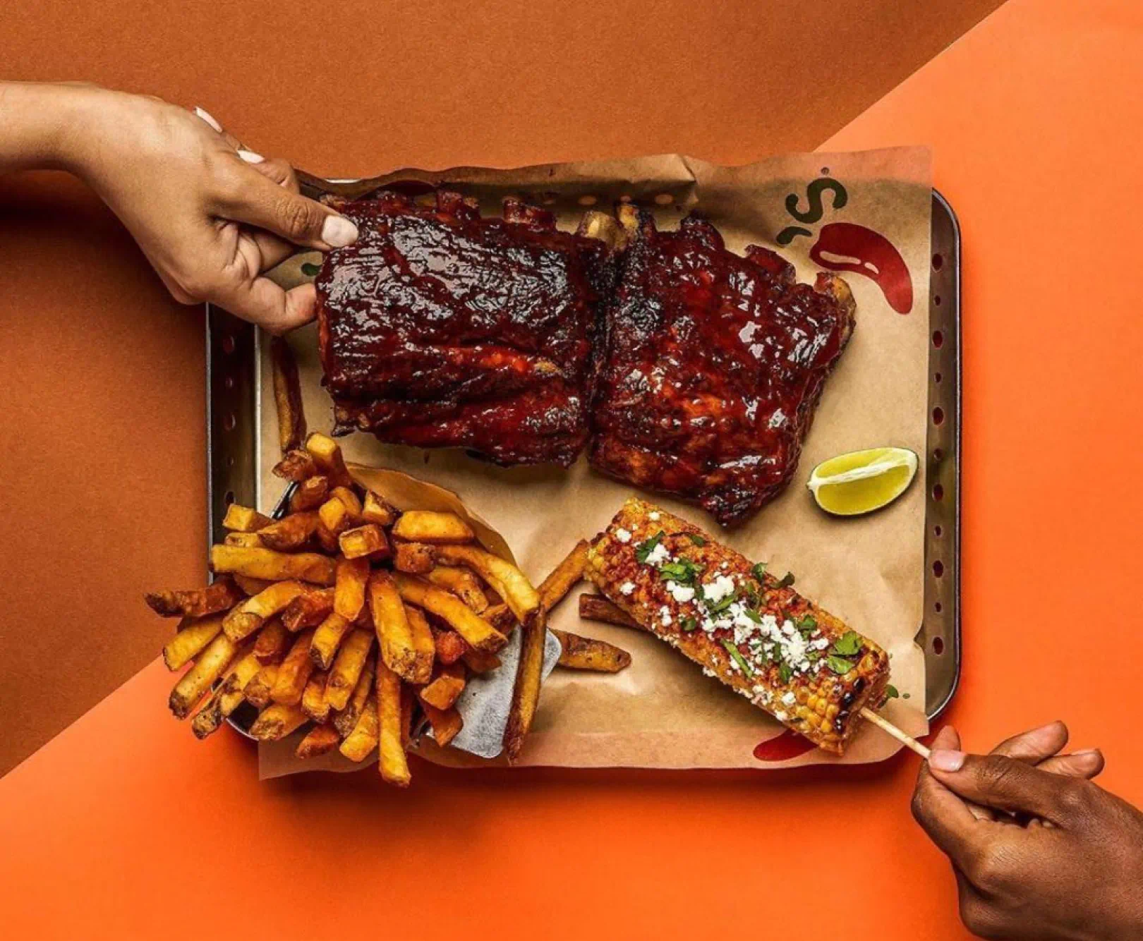 Tray with glazed ribs, seasoned fries, and corn on the cob, against an orange background. Two hands are holding the ribs and corn.