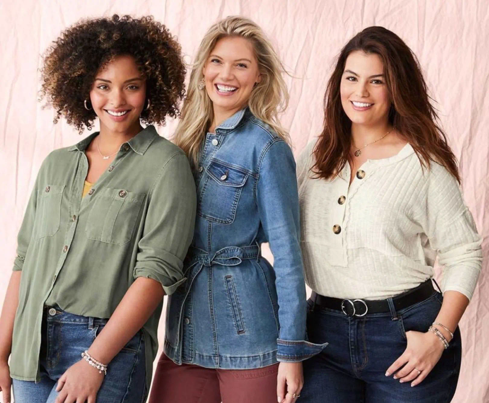 Three women smiling at the camera, dressed casually in button-up shirts and jeans against a light pink backdrop.