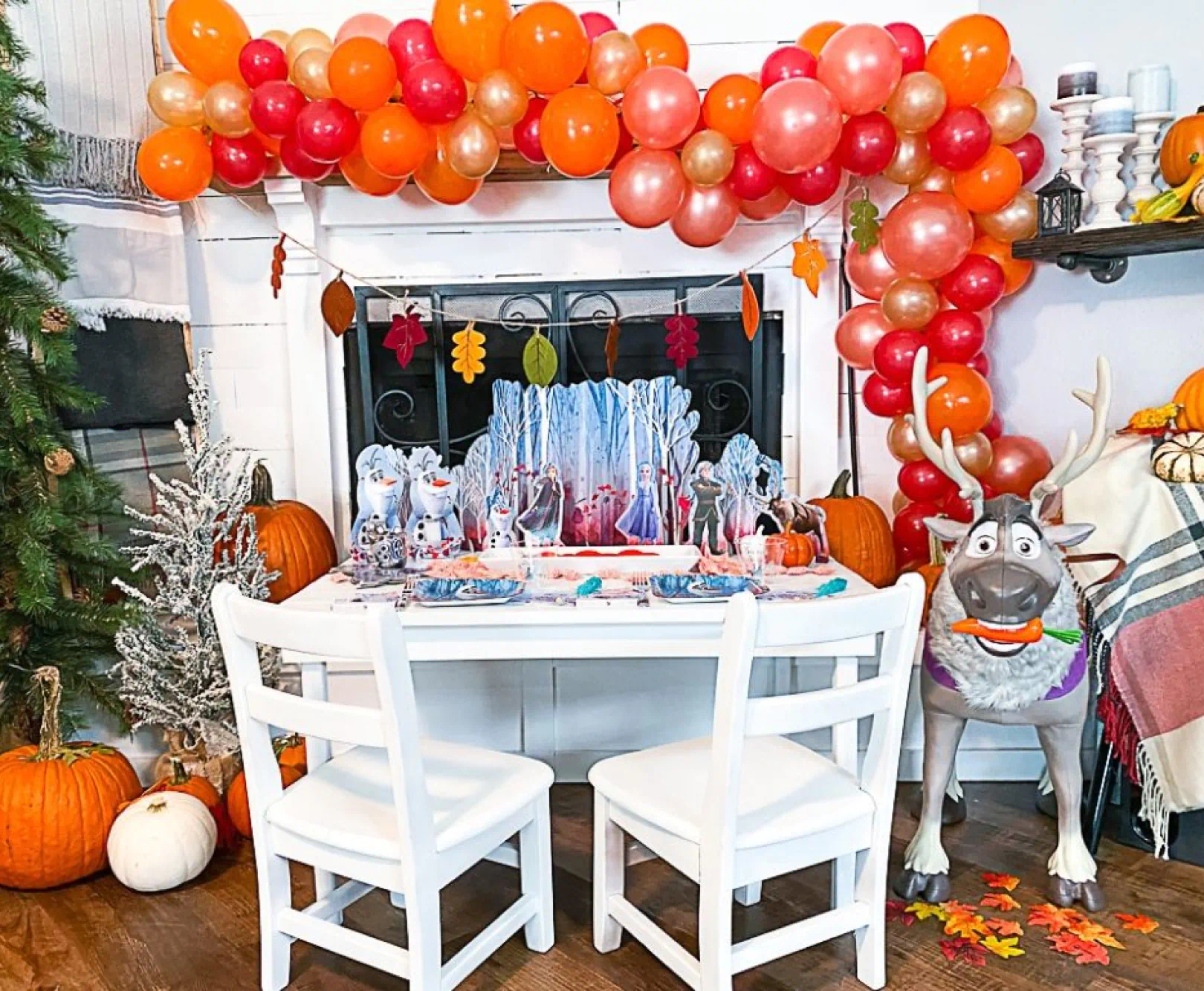 Festive table setup with a white table and chairs, balloon garland, fireplace with leaf decorations, pumpkins, holiday decor, and a stuffed reindeer.