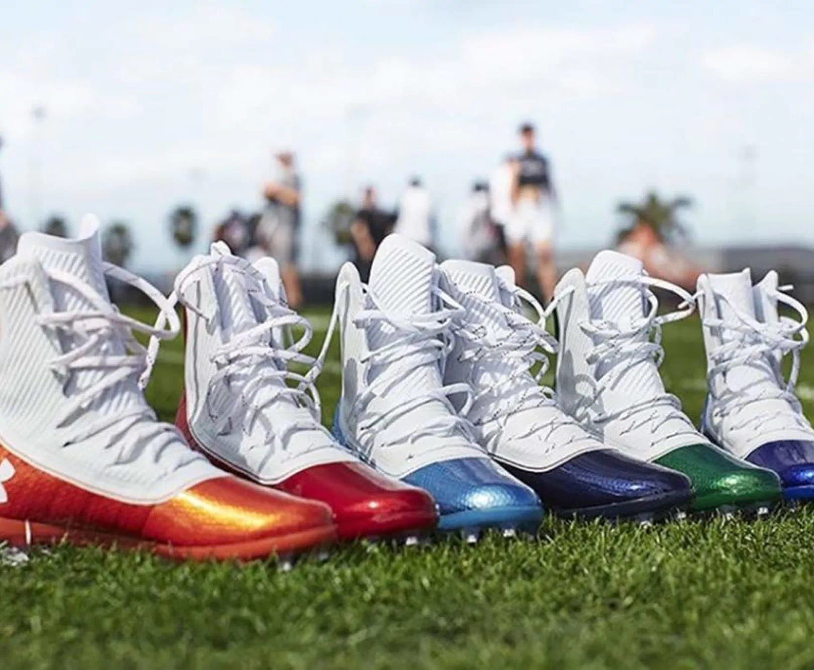 Five football cleats with differently colored soles (orange, red, blue, dark purple, and green) lined up on a grassy field.