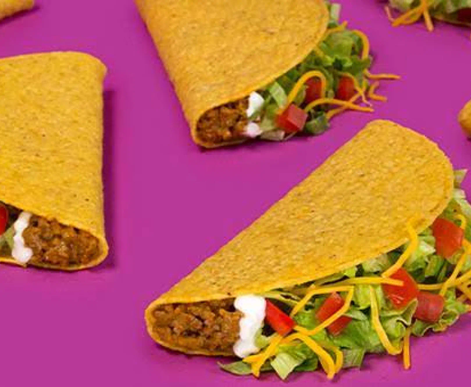 Hard-shell tacos with ground beef, lettuce, tomatoes, cheese, and sour cream on a purple background.