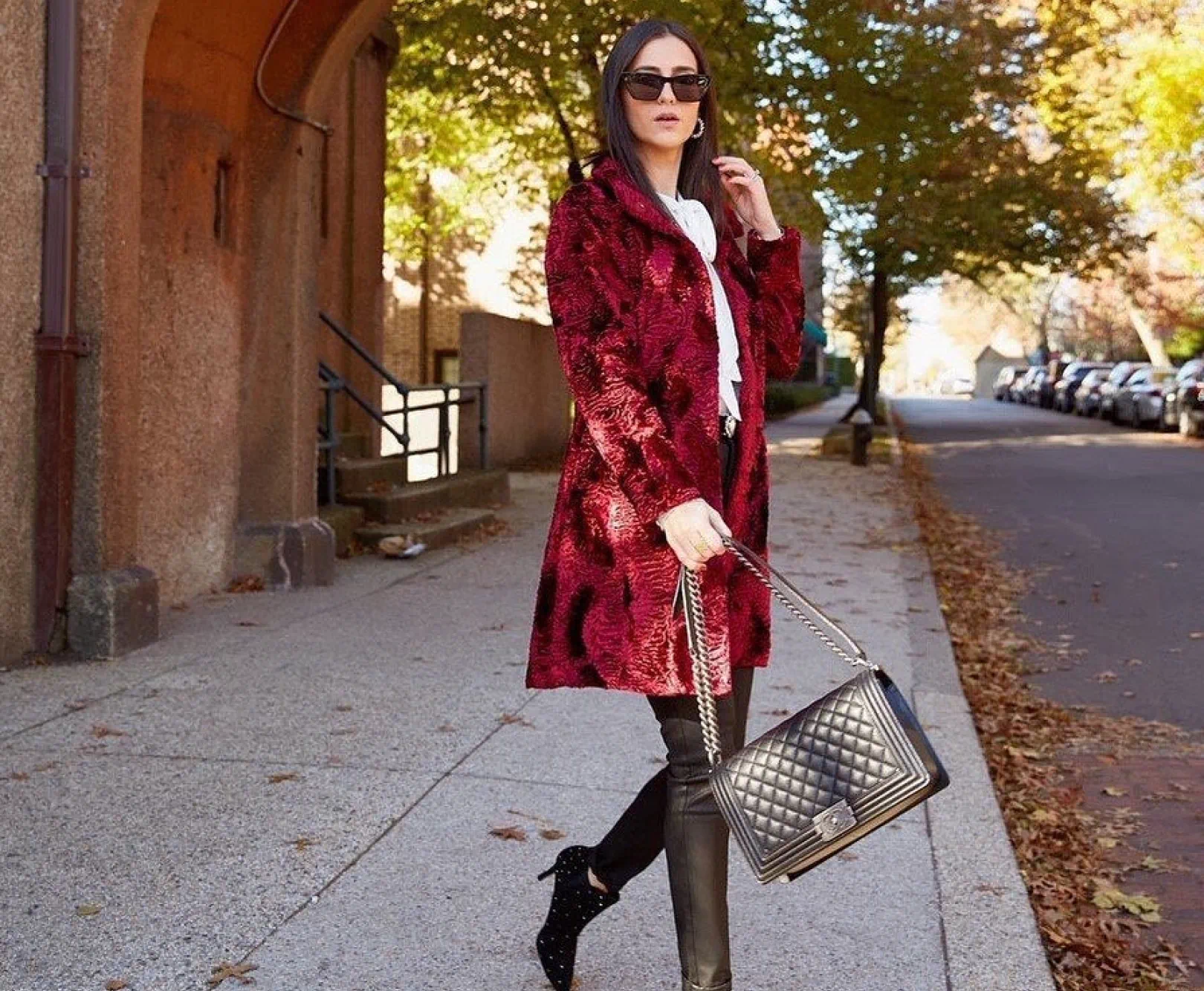 A woman in a red long coat and black sunglasses walking on a sidewalk in an urban area with autumn foliage.