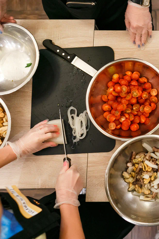 Overhead view of a prep table with silver bowls of sliced cherry tomatoes and prepared sea food, with a woman slicing squid and preparing a meal.