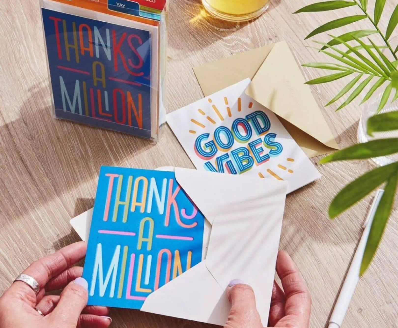 Hands holding a colorful card that says "THANKS A MILLION" next to another card that says "GOOD VIBES" on a wooden table.