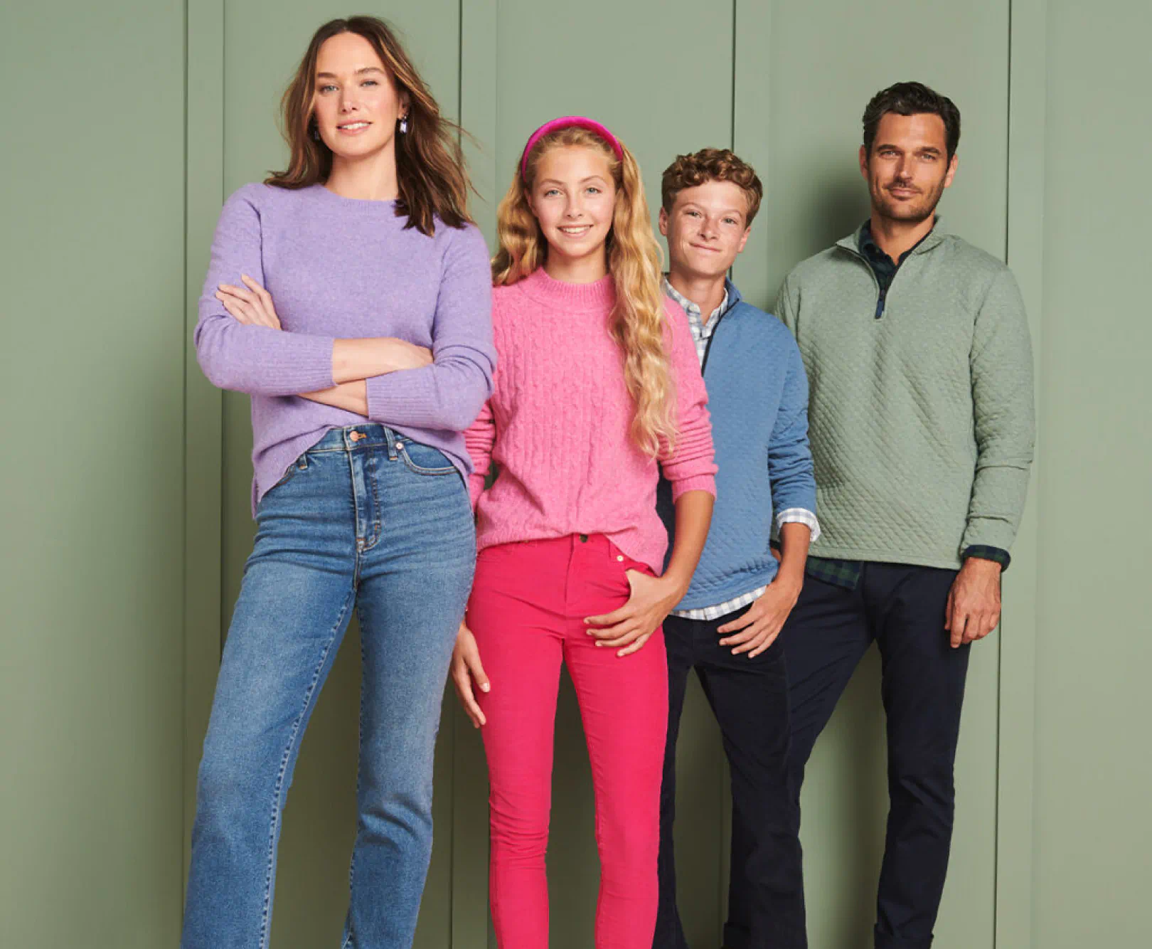 Four people, two adults and two children, standing in front of a muted green, paneled backdrop, wearing colorful casual clothing.