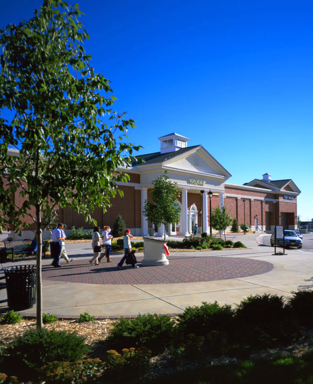 Exterior of a "Von Maur" with people walking through the open space with landscaping in front of the store.