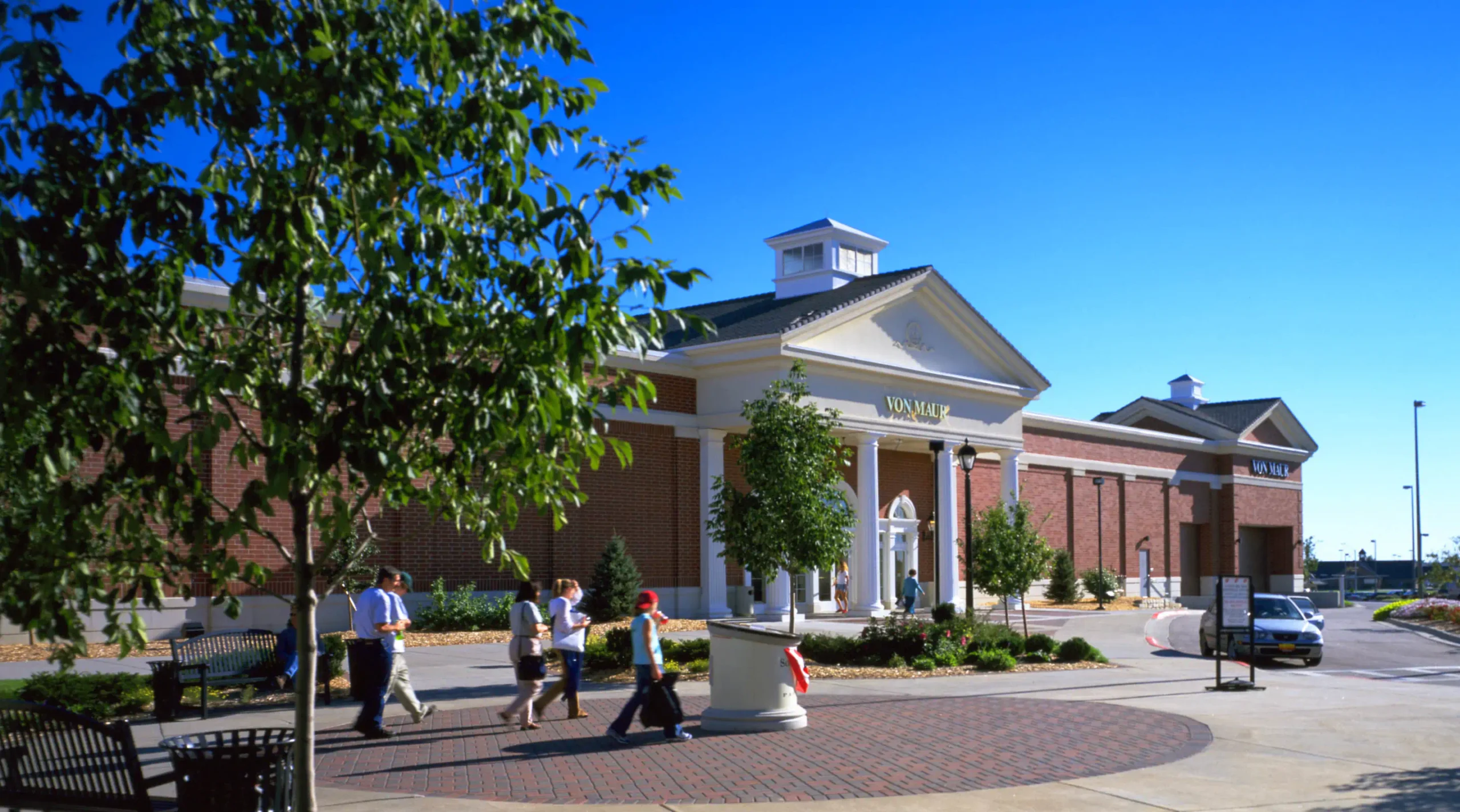 Exterior of a "Von Maur" with people walking through the open space with landscaping in front of the store.