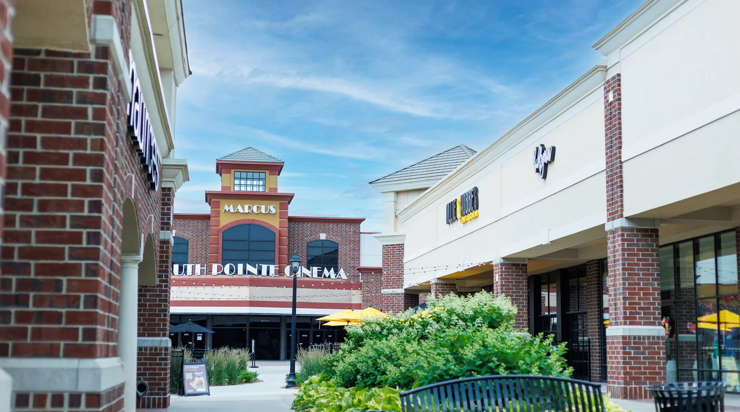 View of the outdoor pavilion at South Pointe, store fronts line the walkway with a bench and landscaping. A cinema sits at the end of the open space.