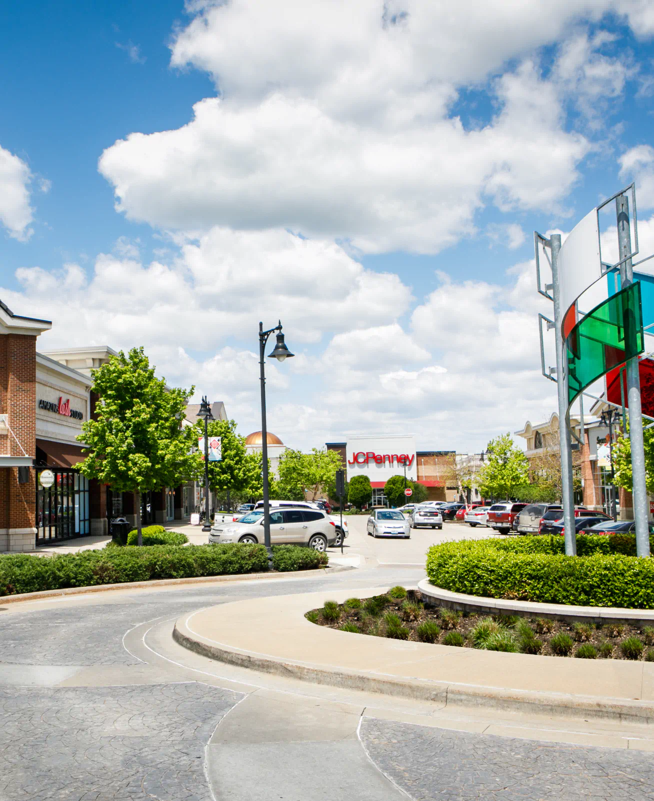 Round about in front of the Summit Fair shops. Store fronts display the signs "Heavenly" and "JCPenney". The roundabout contains landscaping and a colorful metal and glass structure.