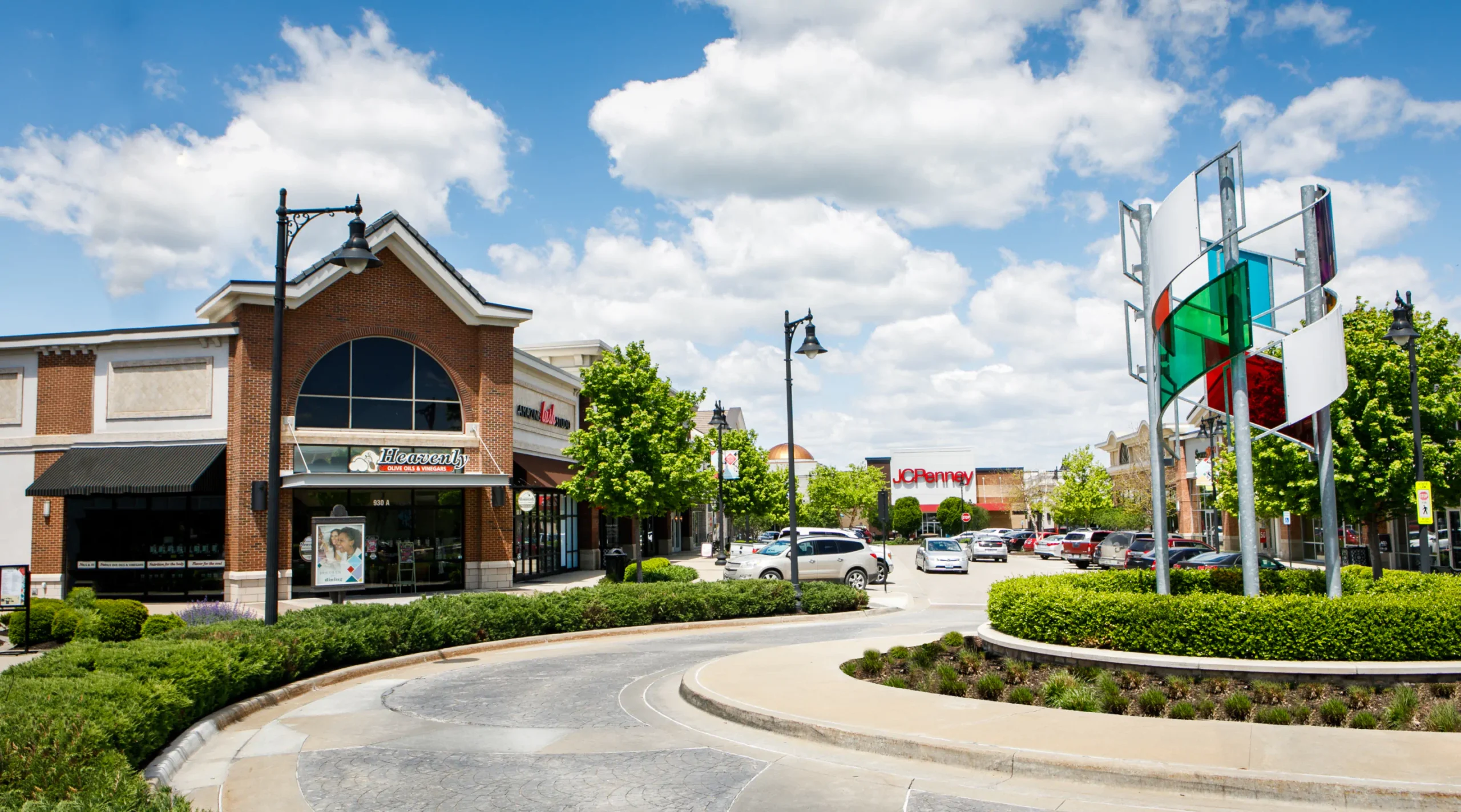 Round about in front of the Summit Fair shops. Store fronts display the signs "Heavenly" and "JCPenney". The roundabout contains landscaping and a colorful metal and glass structure.