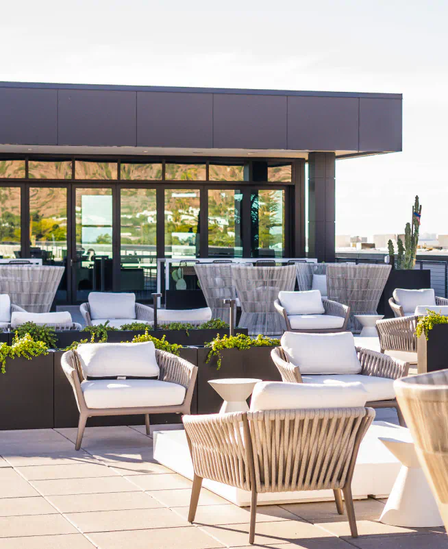 Outdoor rooftop lounge area with modern furniture, and a dark grey building with large glass windows in the background.