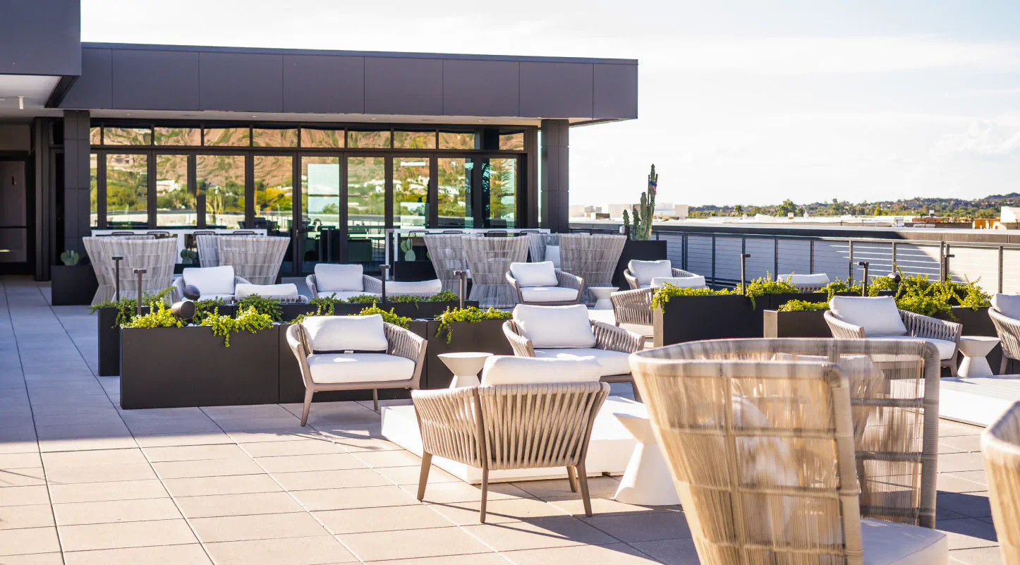 Modern rooftop patio with white cushioned seating, black planters with greenery, and large glass doors in the background.