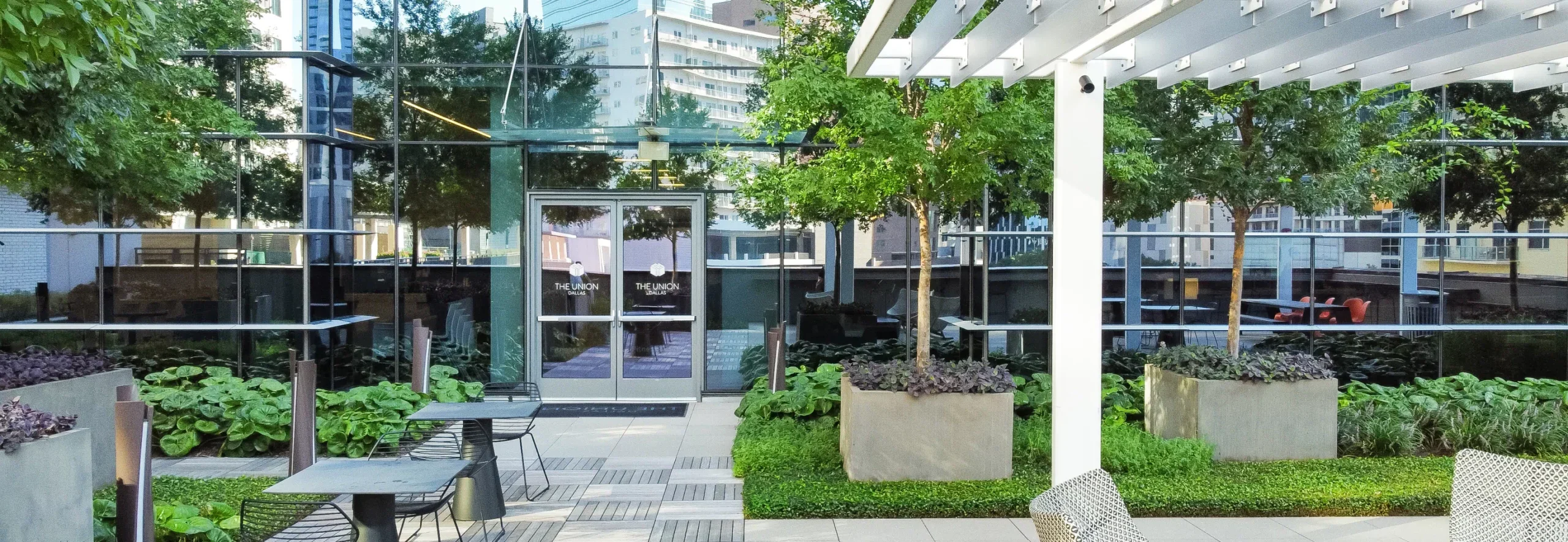 Entrance to The Union, "The Union" logo printed on double glass doors to a glass building. In front of the doors is an outdoor patio space with places to sit, landscaping, trees and plants.