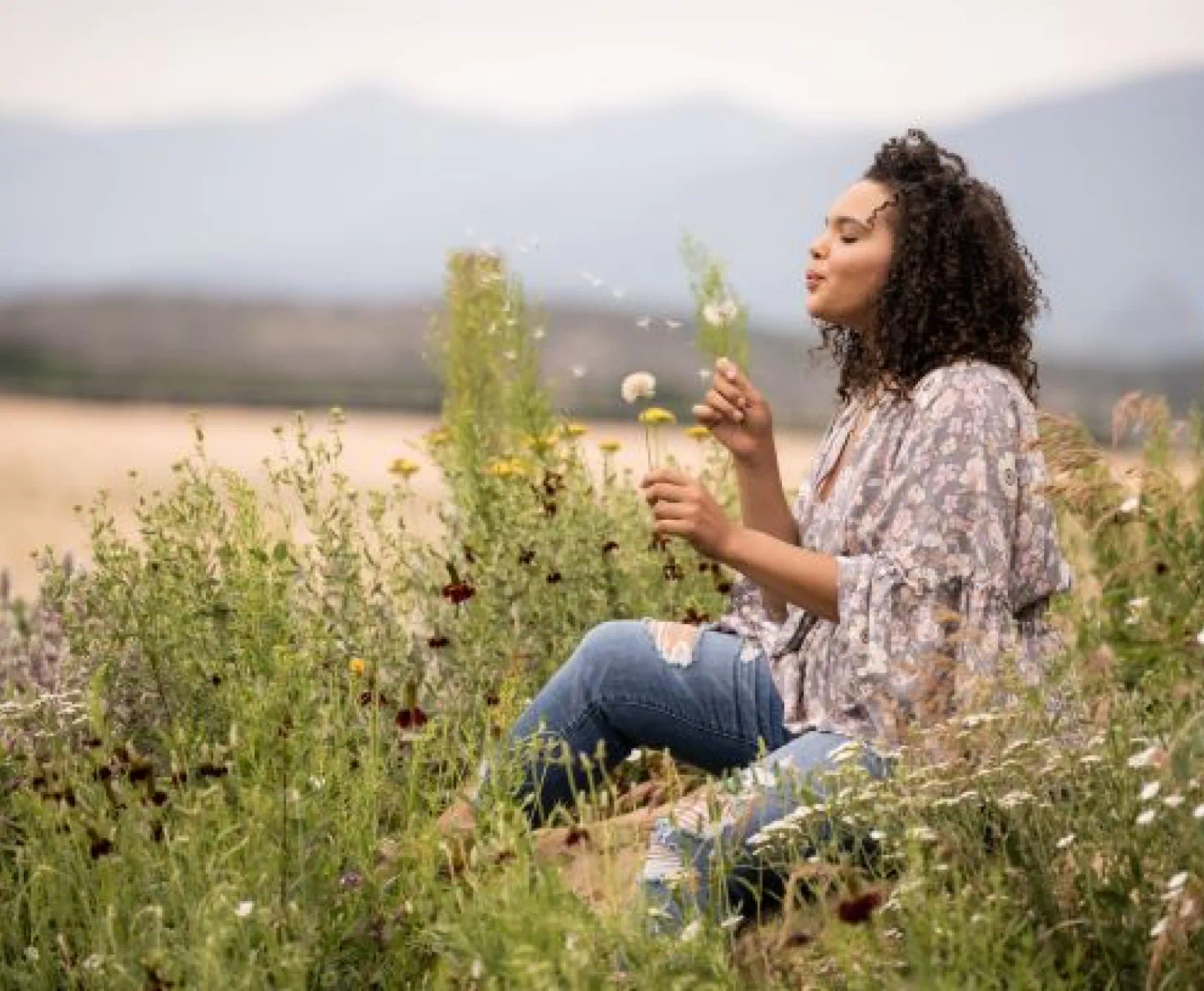 A woman sits in a field blowing the seeds off a dandelion.