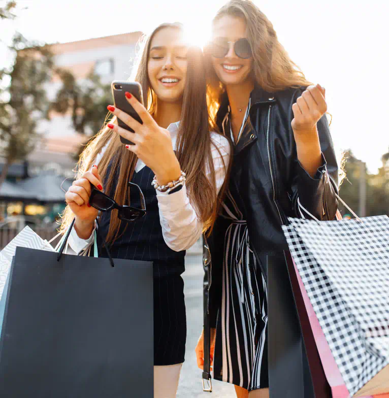 Two fashionably dressed people holding shopping bags smile at a phone.