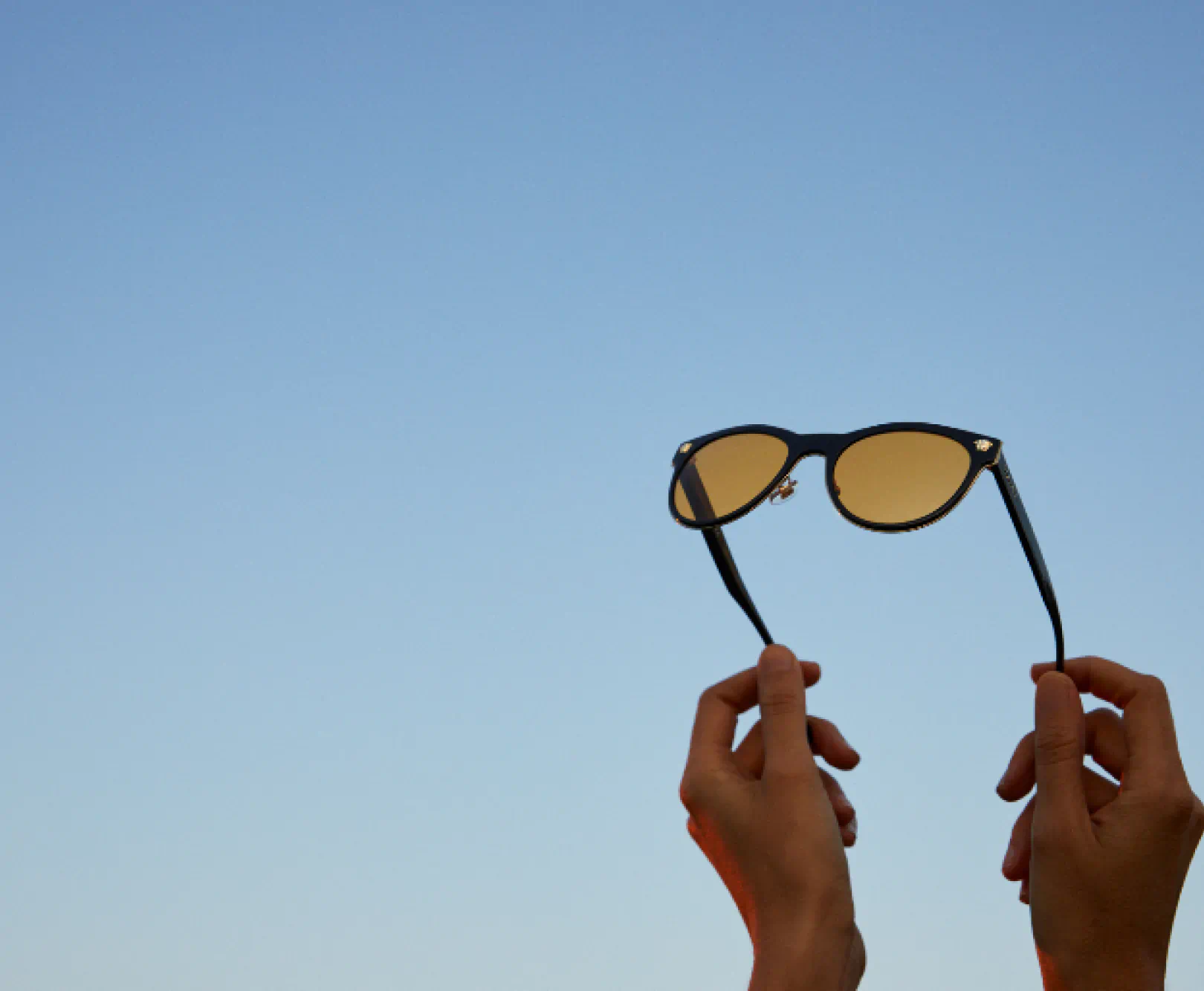 A person holds a pair of sunglasses towards a cloudless sky. The frames of the glasses are black, the lenses are brown.