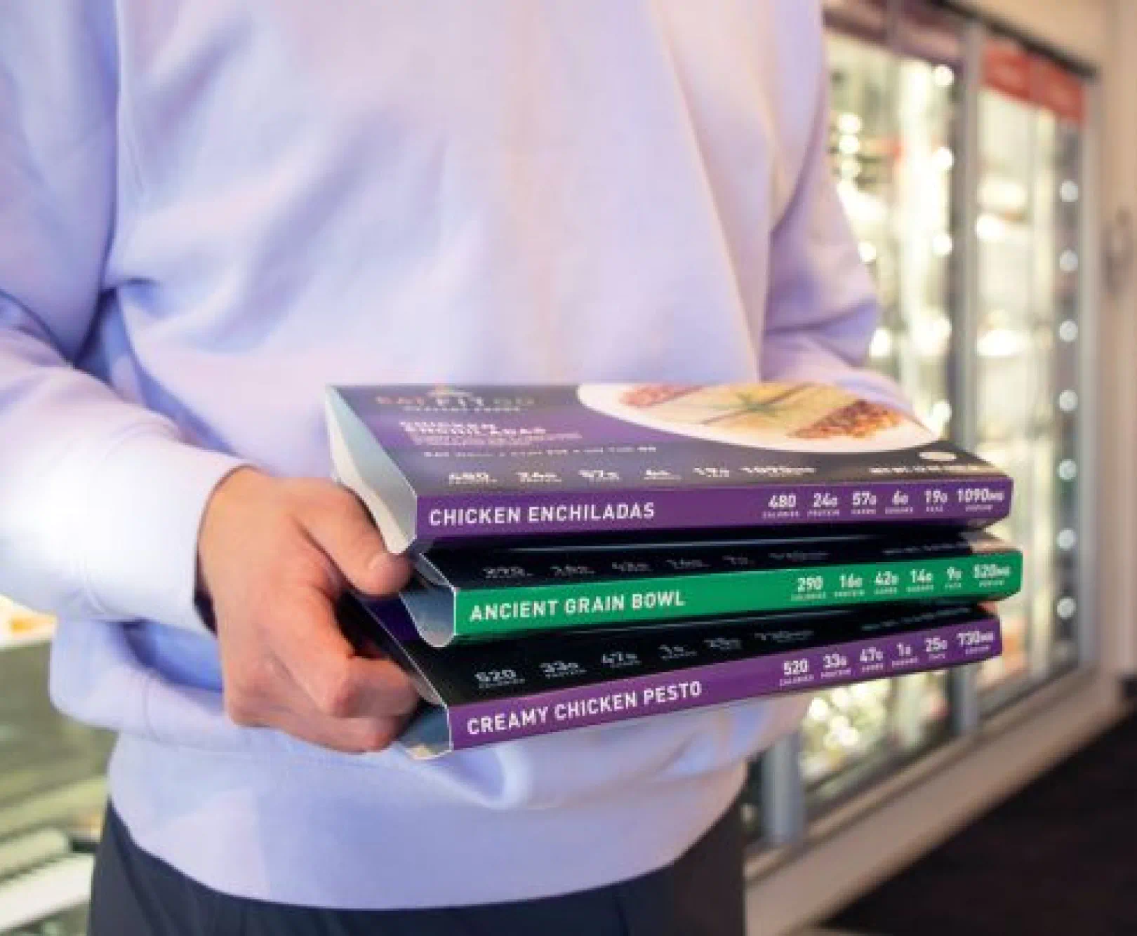 A man in a lavender sweater holds three ready-made meals from Eat Fit Go.