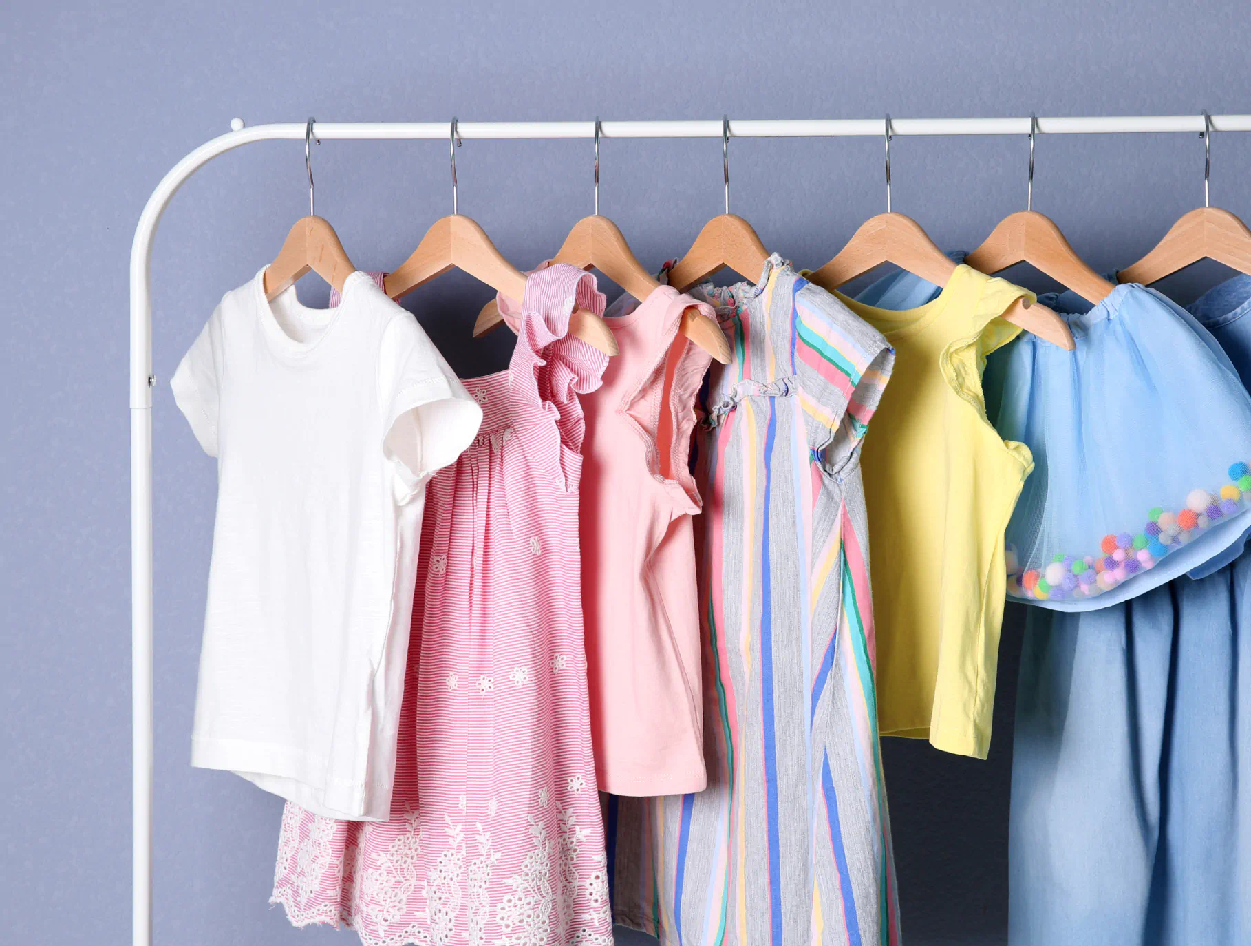 A white clothing rack holds several hangers of kids dresses and tops. The clothing is assorted in white, pink, pastel and rainbow colors.