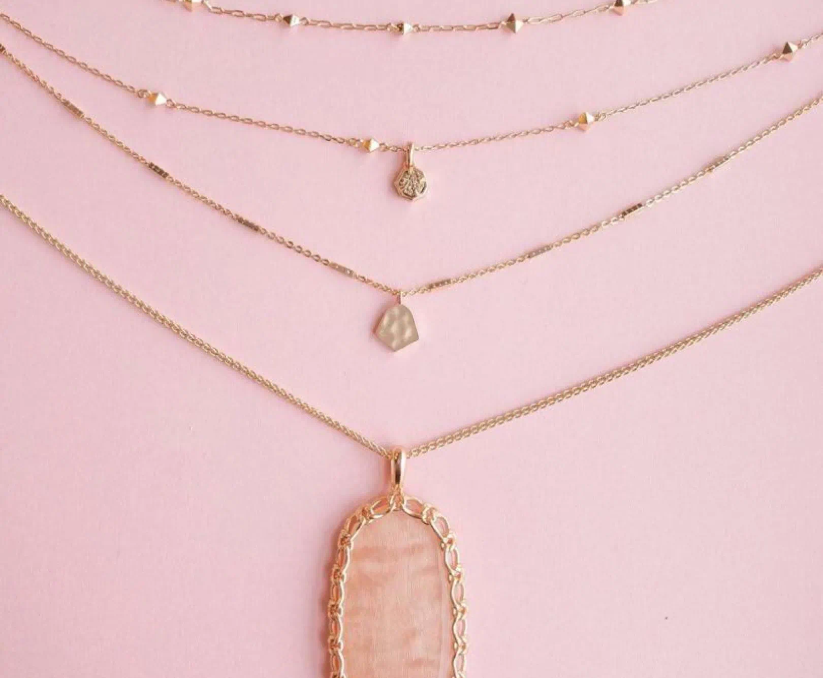 A collection of gold necklaces with pendants lay across a pink tablecloth.