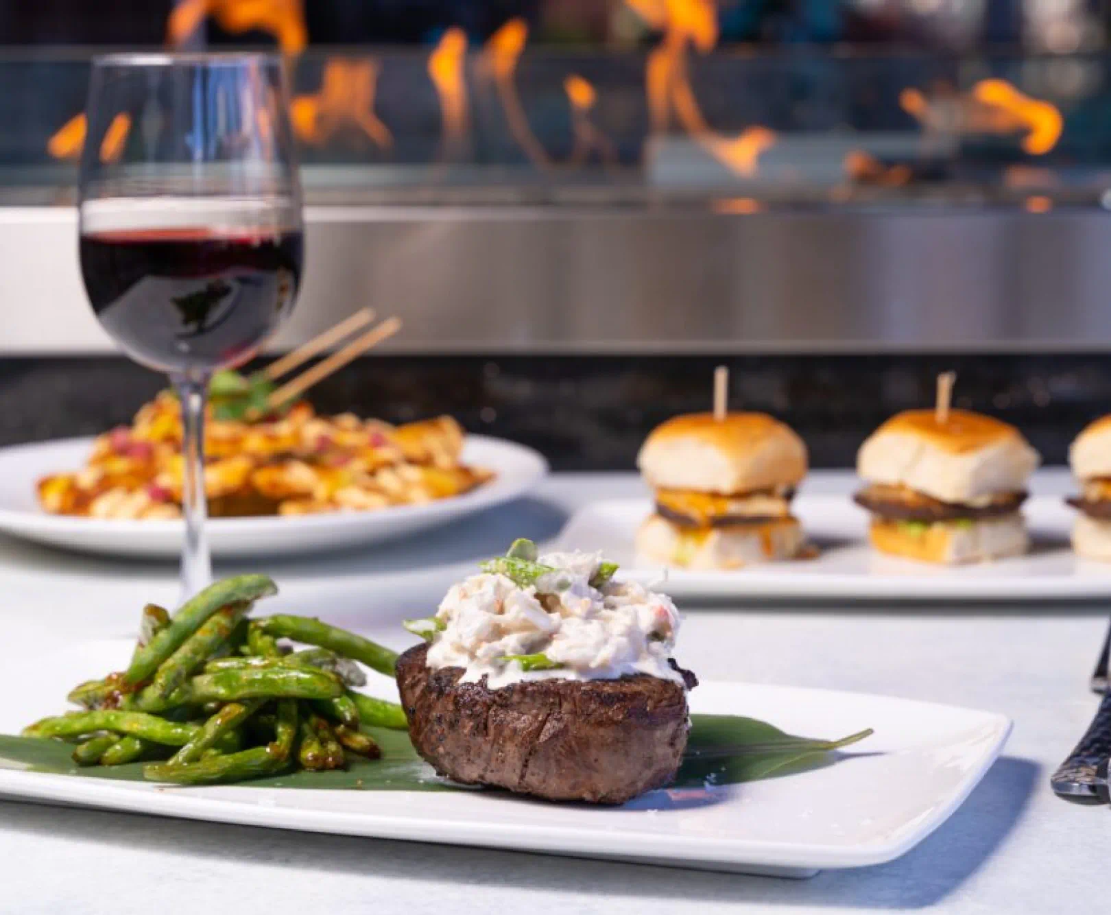 A loaded baked potato sits on a white plate, served with asparagus and red wine. Some sliders and a pasta can been seen in the background.