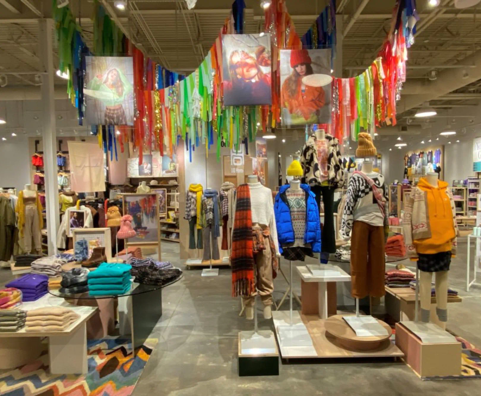 The indoor of fully stocked clothing store features bright and colorful winter clothing on shelves. Rainbow-colored streamers and metallic tinsel hang from the ceiling.